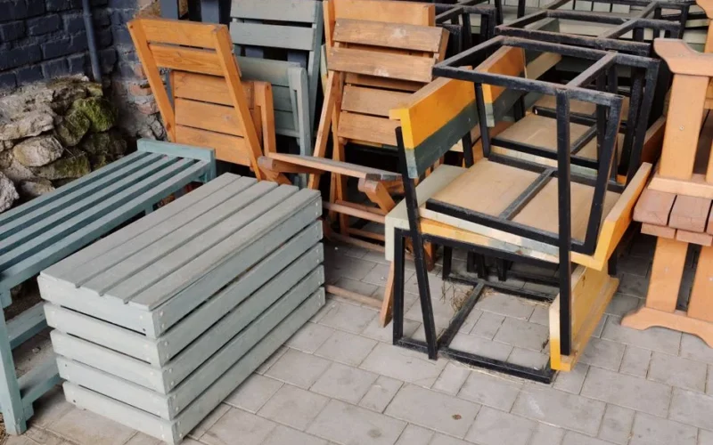 A mix of wooden benches, chairs, and metal frames stacked outside on a paved area.