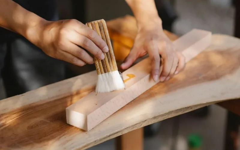 High angle of crop anonymous male artisan oiling wooden plank with brush while working in workshop