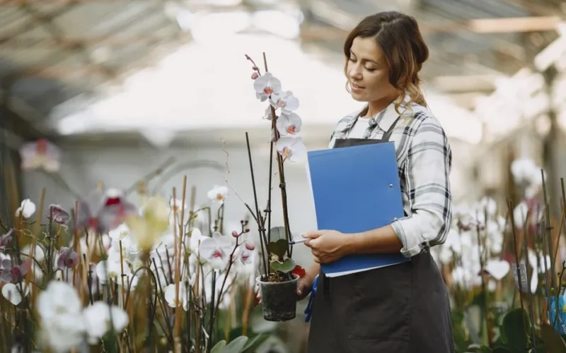Woman in greenhouse holding a potted orchid and clipboard, surrounded by flowers.