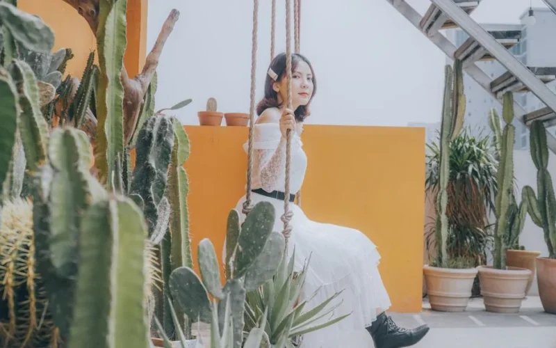 A woman in a white dress sits on a swing surrounded by cacti, creating a vibrant urban garden scene.