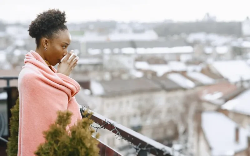 A woman wrapped in a blanket enjoys a hot drink on a chilly city balcony.