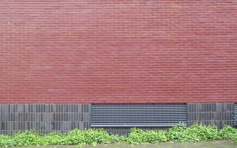 A minimalist photo of a red brick wall featuring green ivy and a ventilation grille.