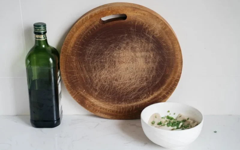 A rustic kitchen setup featuring dumplings in a bowl and an oil bottle next to a wooden cutting board.