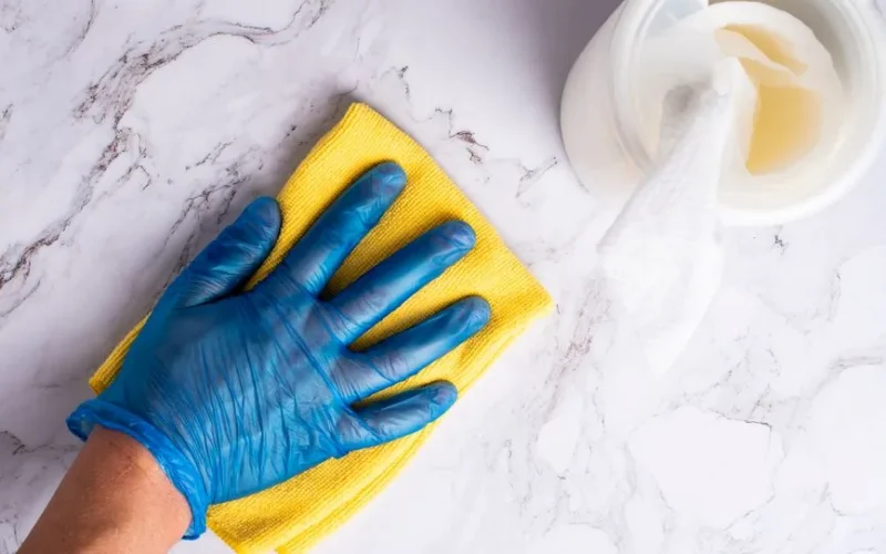 A gloved hand cleans a marble countertop with a yellow cloth next to disinfectant wipes.