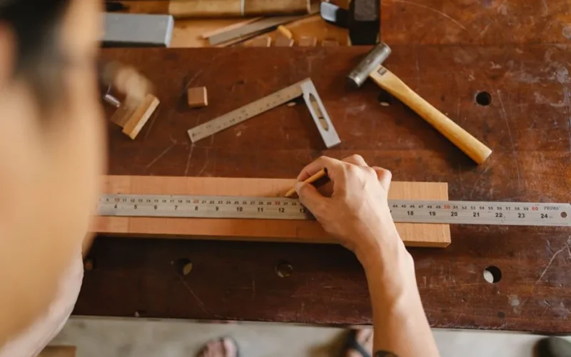 Top view of crop anonymous male measuring timber plank with ruler and marking with pencil while working on table with tools