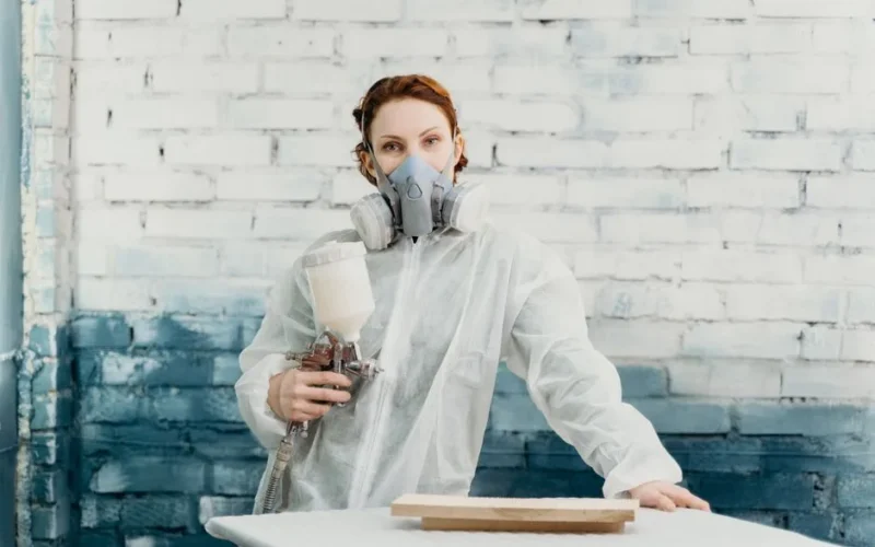 Woman in protective suit and gas mask using spray gun indoors against a painted brick wall.