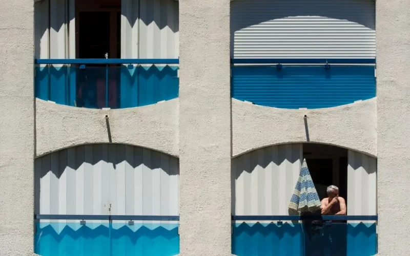 A man stands on a blue-themed balcony under sunlight, enjoying a summer day.