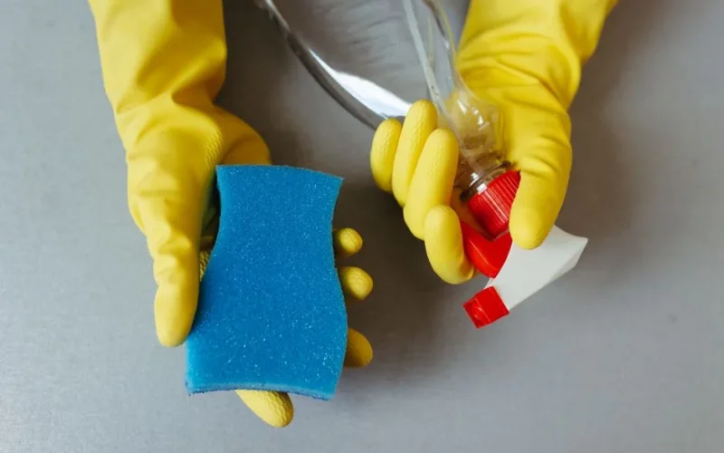 Hands wearing yellow gloves holding a sponge and a spray bottle.