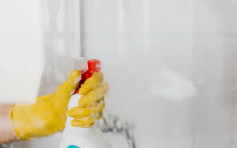 Crop unrecognizable person in yellow latex protective glove spraying detergent from bottle with plastic dispenser at glass of shower cabin