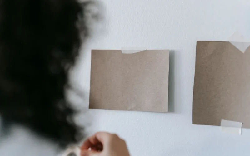 Woman hangs blank cardboard pieces on a wall using adhesive tape, indoors.