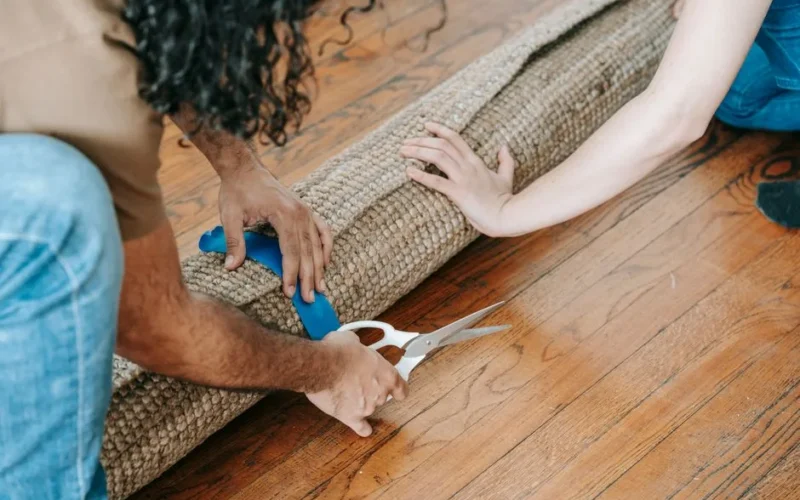 Two adults preparing to move by rolling a carpet on wooden flooring indoors.