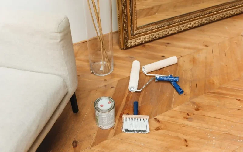 Paint supplies laid out on a wooden floor beside a couch and a large framed mirror, ready for home renovation.