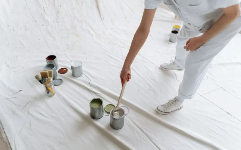 Person in white attire painting on a white surface with various color paint cans and brushes.