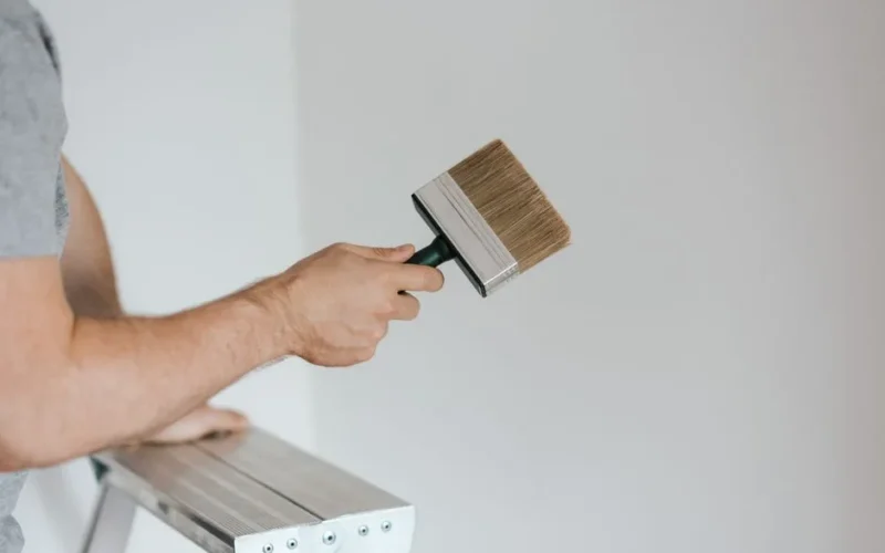 Side view of crop faceless painter in casual clothes holding paintbrush while leaning on stepladder on gray background in soft daylight