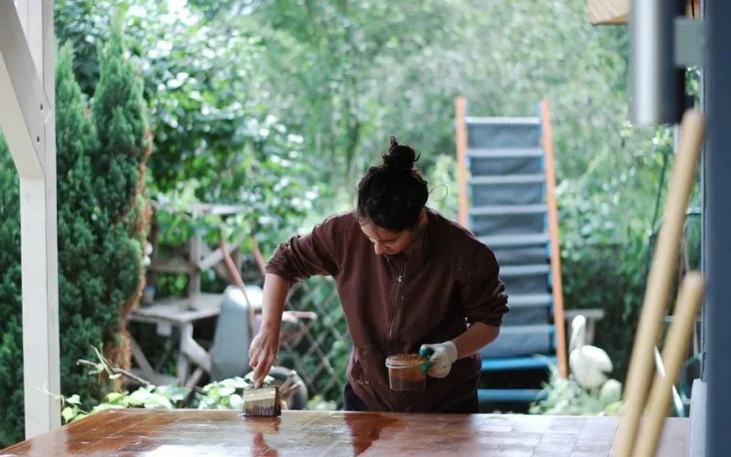 Woman applies varnish to a table outdoors, surrounded by greenery. DIY and home improvement scene.