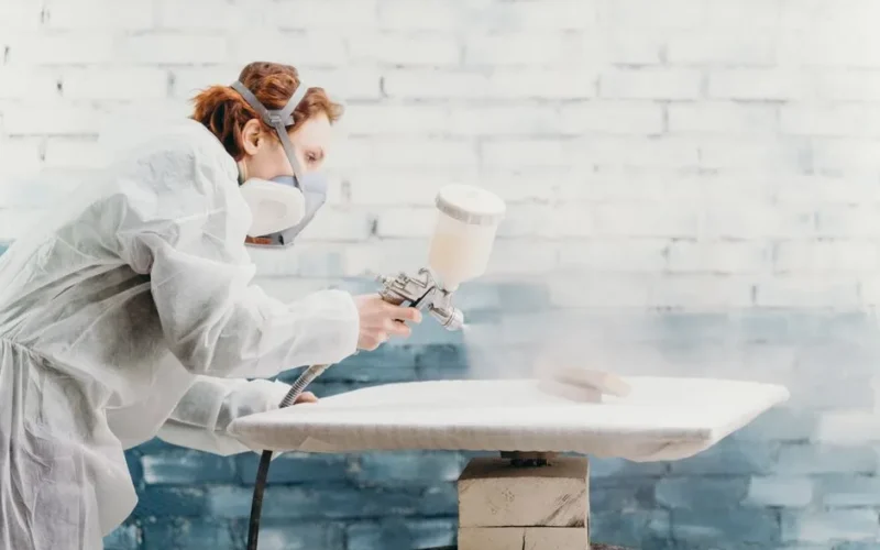 Redheaded woman wearing protective gear spray paints a furniture piece indoors.