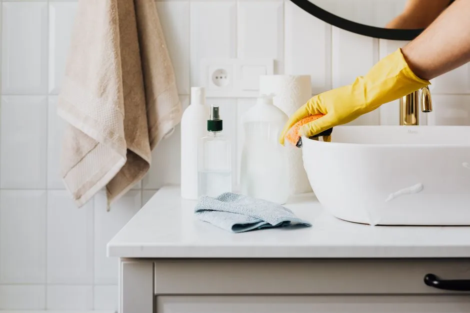 Crop anonymous person in yellow rubber protective glove wiping surface of white stylish sink with sponge and cleaning agent in modern bathroom interior